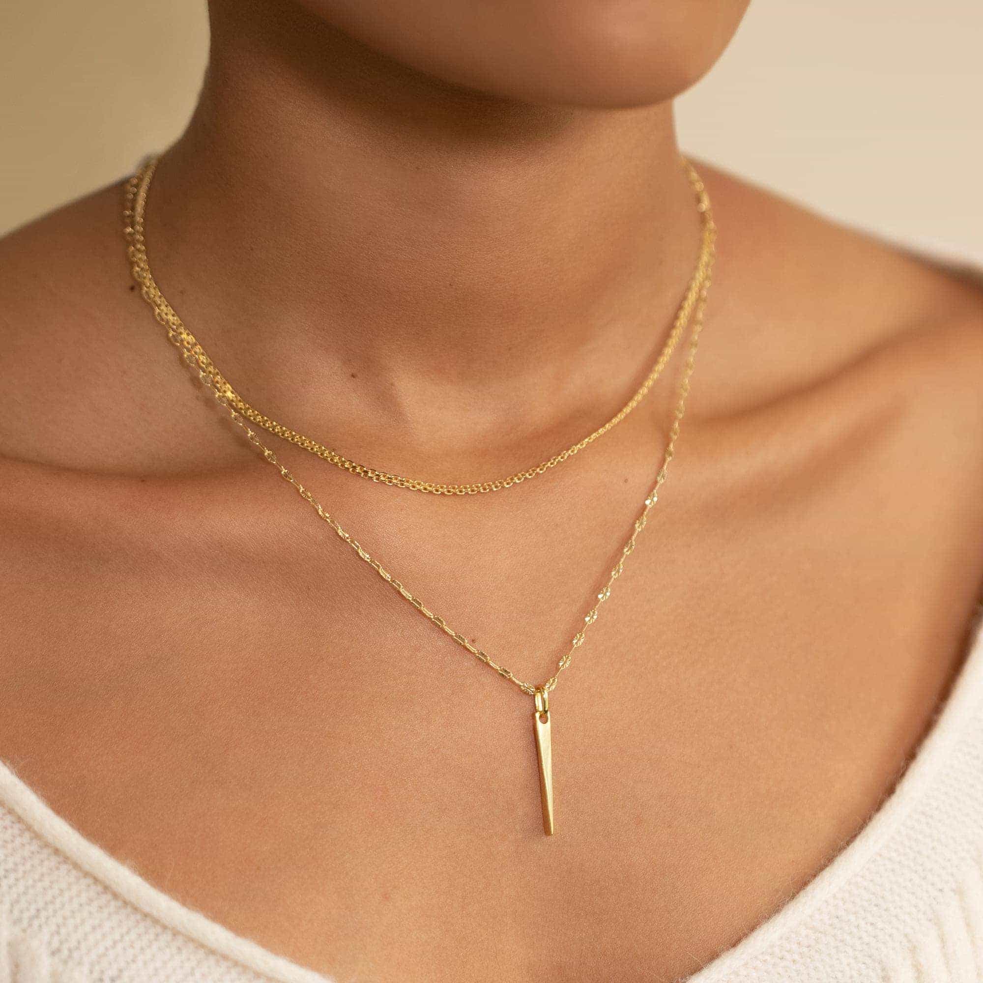 Gold fine chain necklace with a slim spike pendant, shown layered on a model's neckline