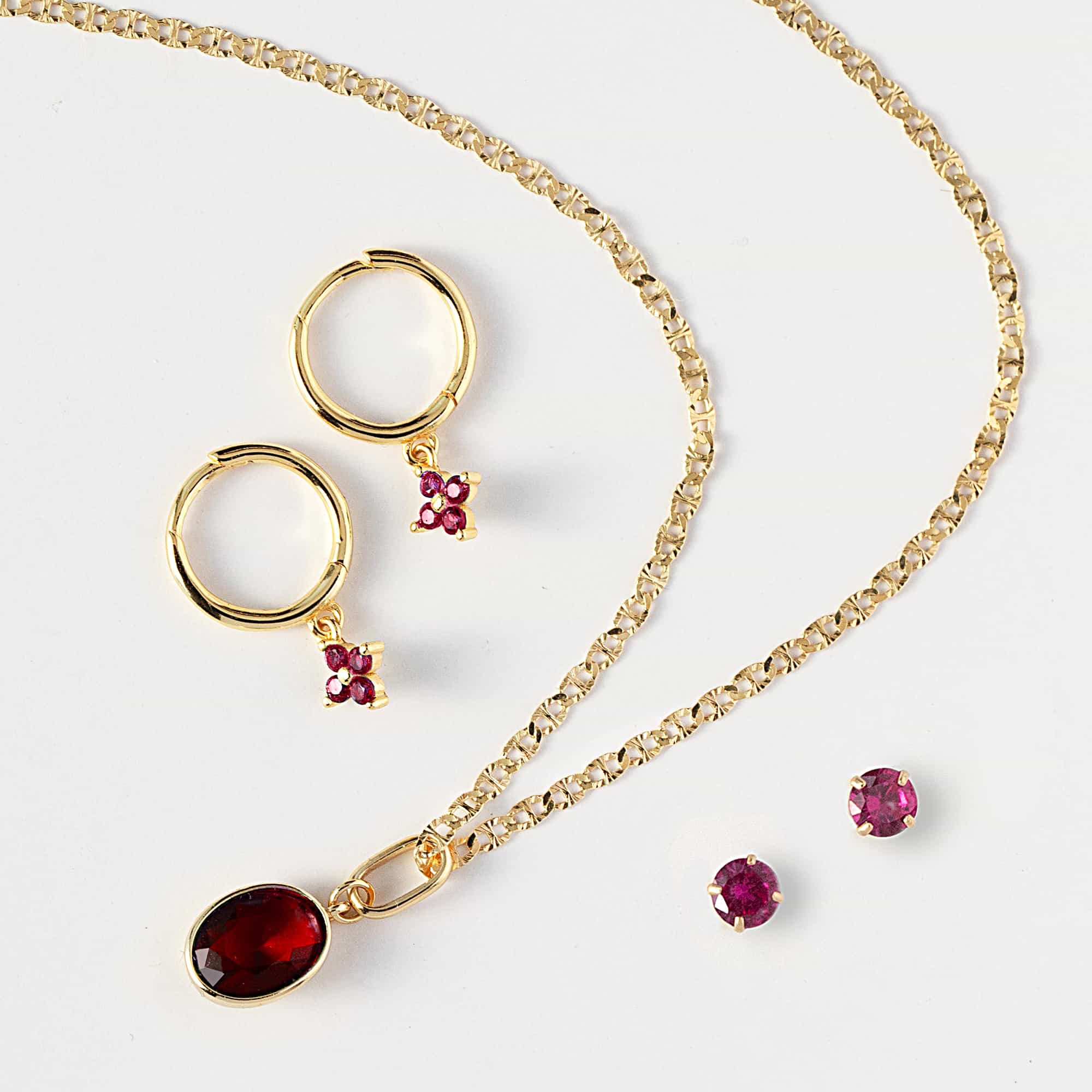 Gold flatlay with charm huggie hoops, a chain, a red-stone pendant and red-stone studs, on a white background