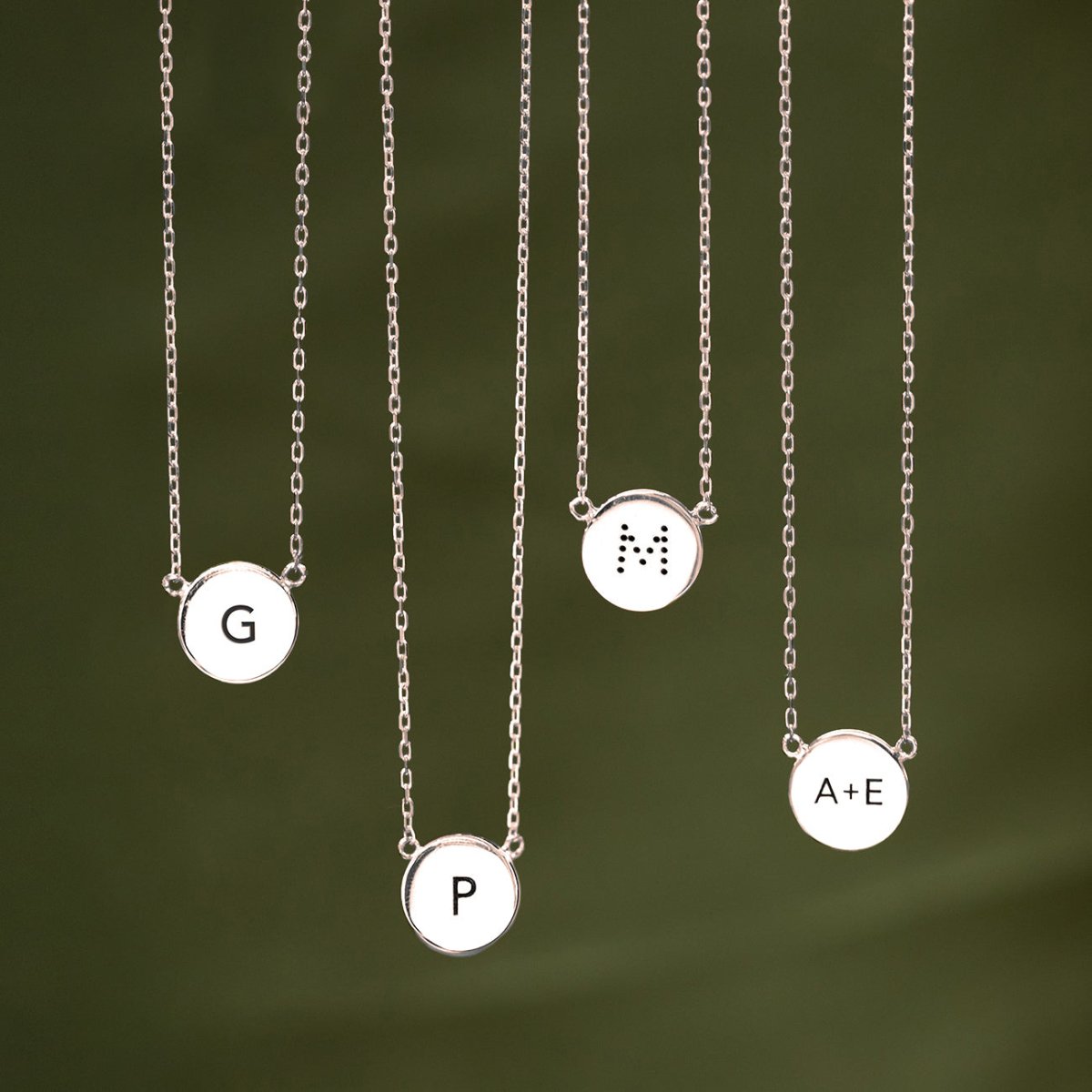Four sterling silver disc necklaces each engraved with an initial, shown against a green background