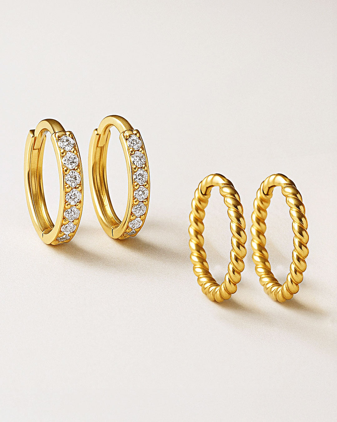 Two pairs of gold huggie hoops on a pale surface, one set with clear stones and one with a twisted rope design