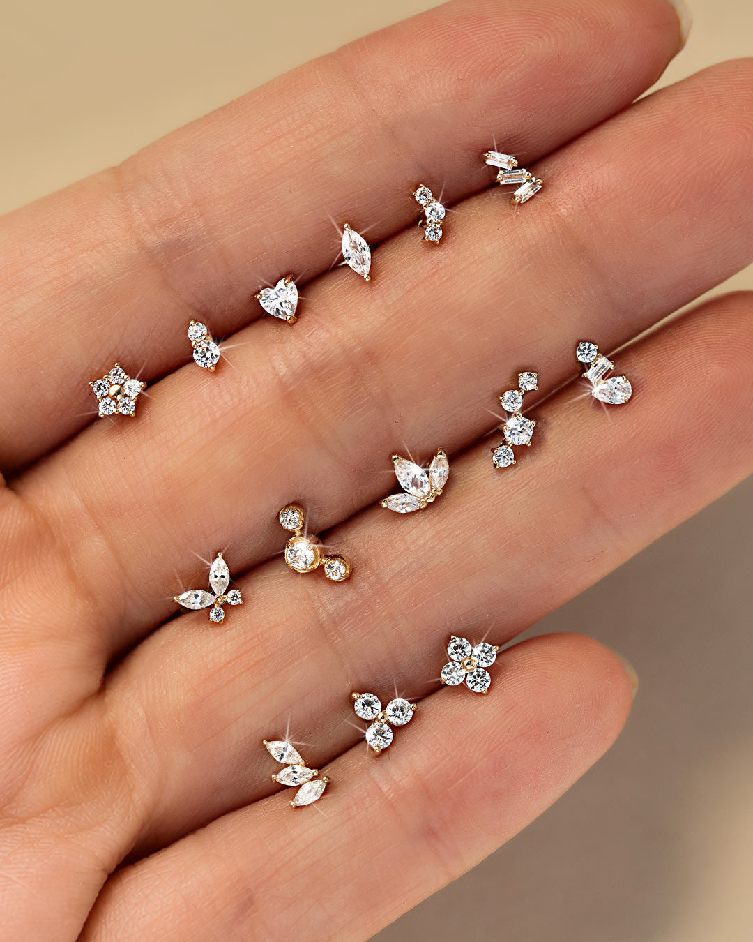 Twelve gold crystal flat back studs in star, heart, marquise, butterfly and cluster shapes displayed between fingers.