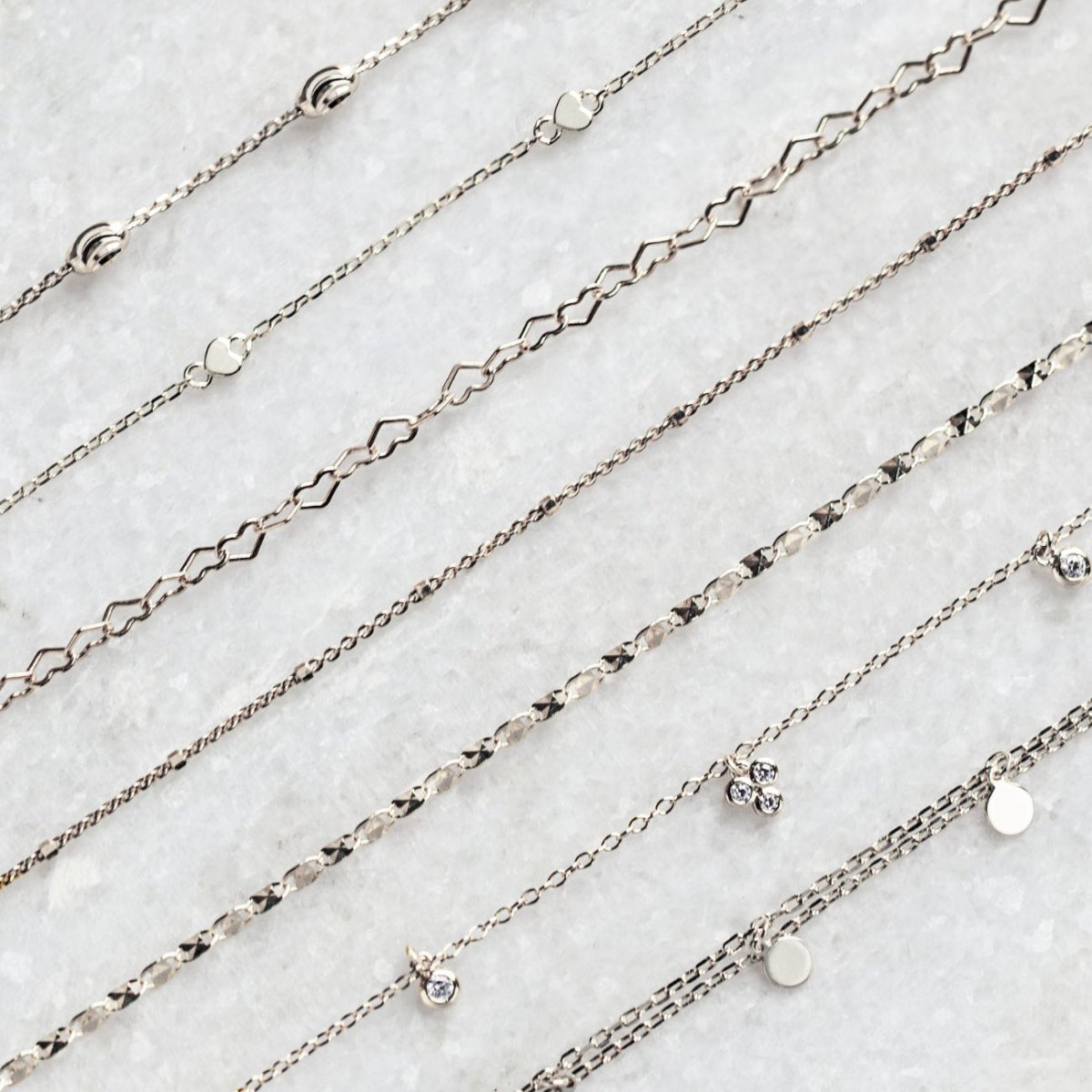 An assortment of dainty silver anklet chains in varied link styles, laid in rows on white marble