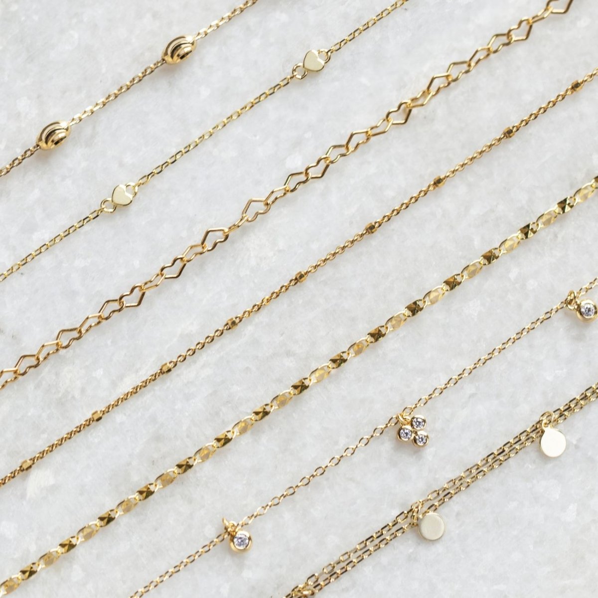 An assortment of dainty gold anklet chains in varied link styles, laid in rows on white marble