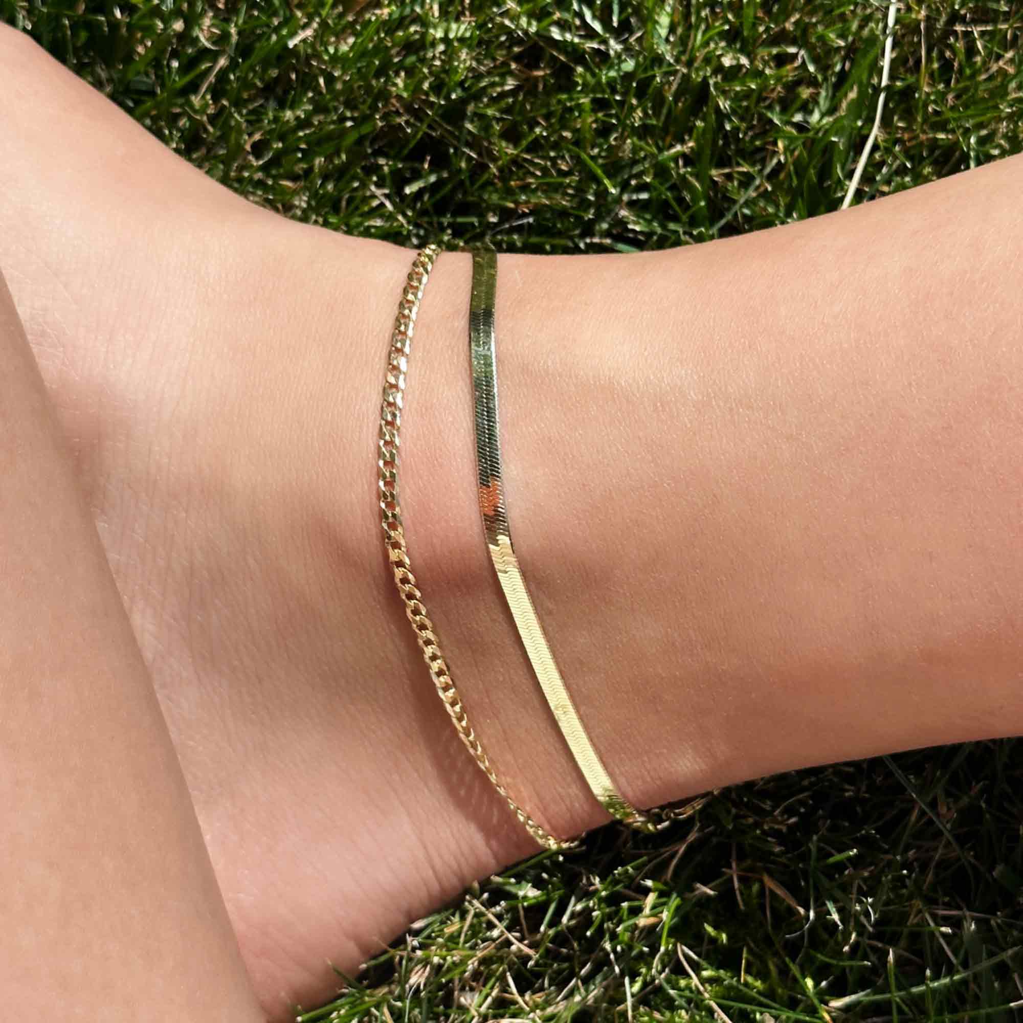 Gold vermeil herringbone chain anklet layered with a curb chain, shown on a model's ankle in grass