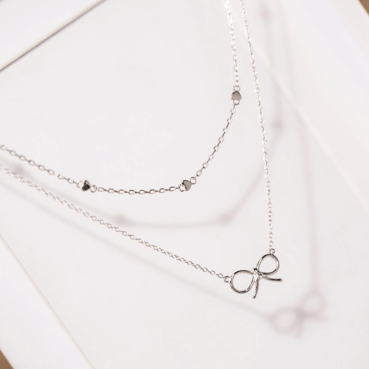 Sterling silver heart-station chain necklace shown layered with a bow pendant chain, on a white background