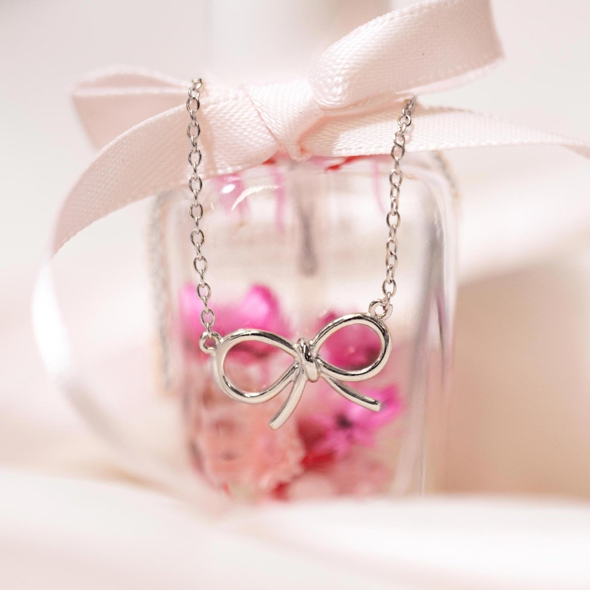 Sterling silver bow necklace draped over a glass bottle with pink ribbon and dried flowers