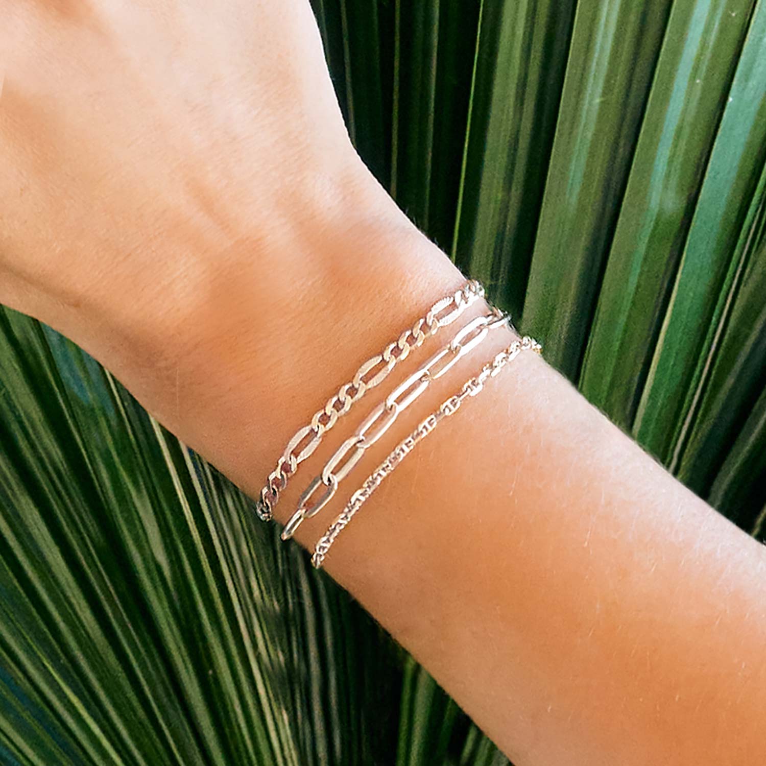 Sterling silver faceted Figaro chain bracelet layered on a model's wrist against palm leaves