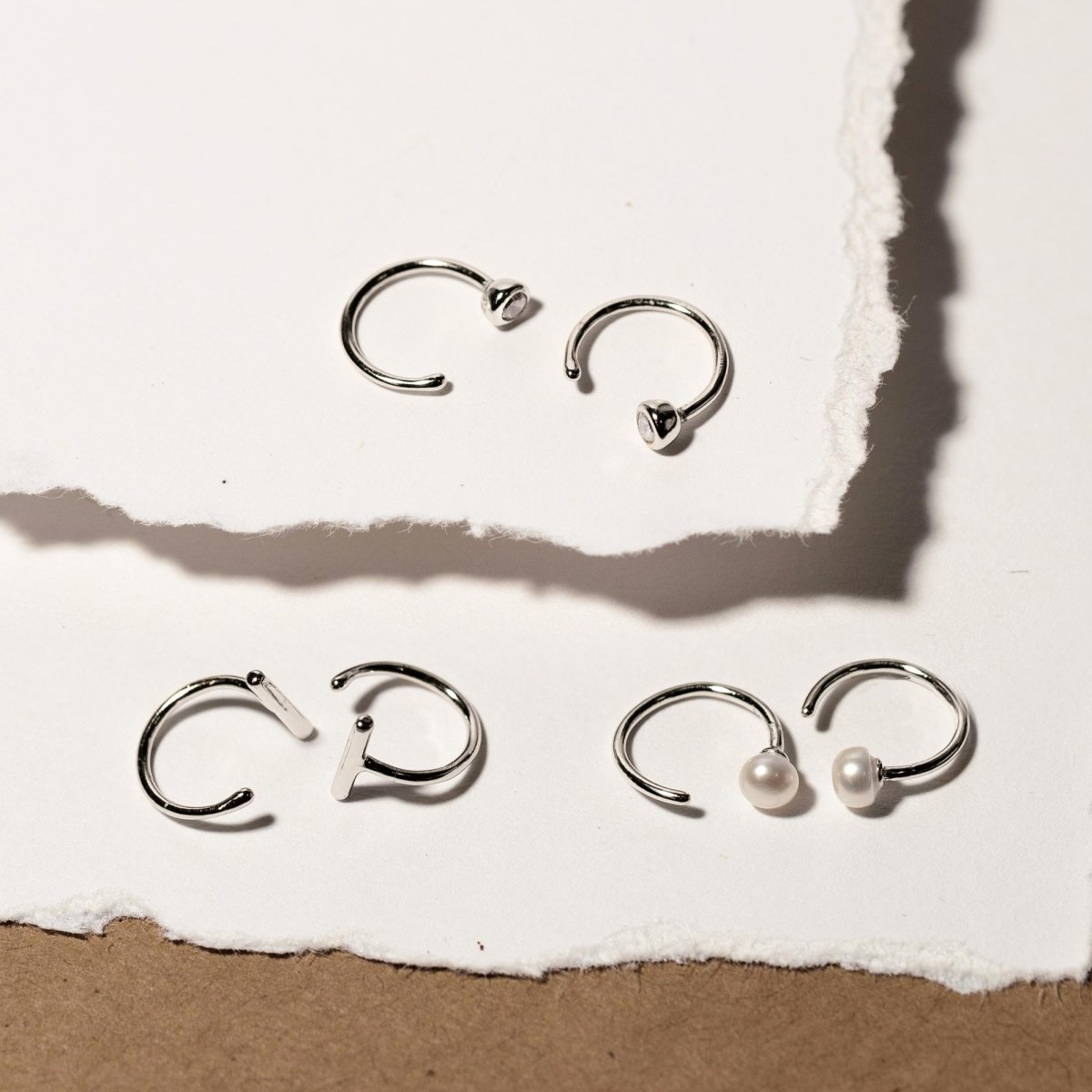 Sterling silver bar huggie earrings with clear stone and pearl variants, flat-laid on torn paper