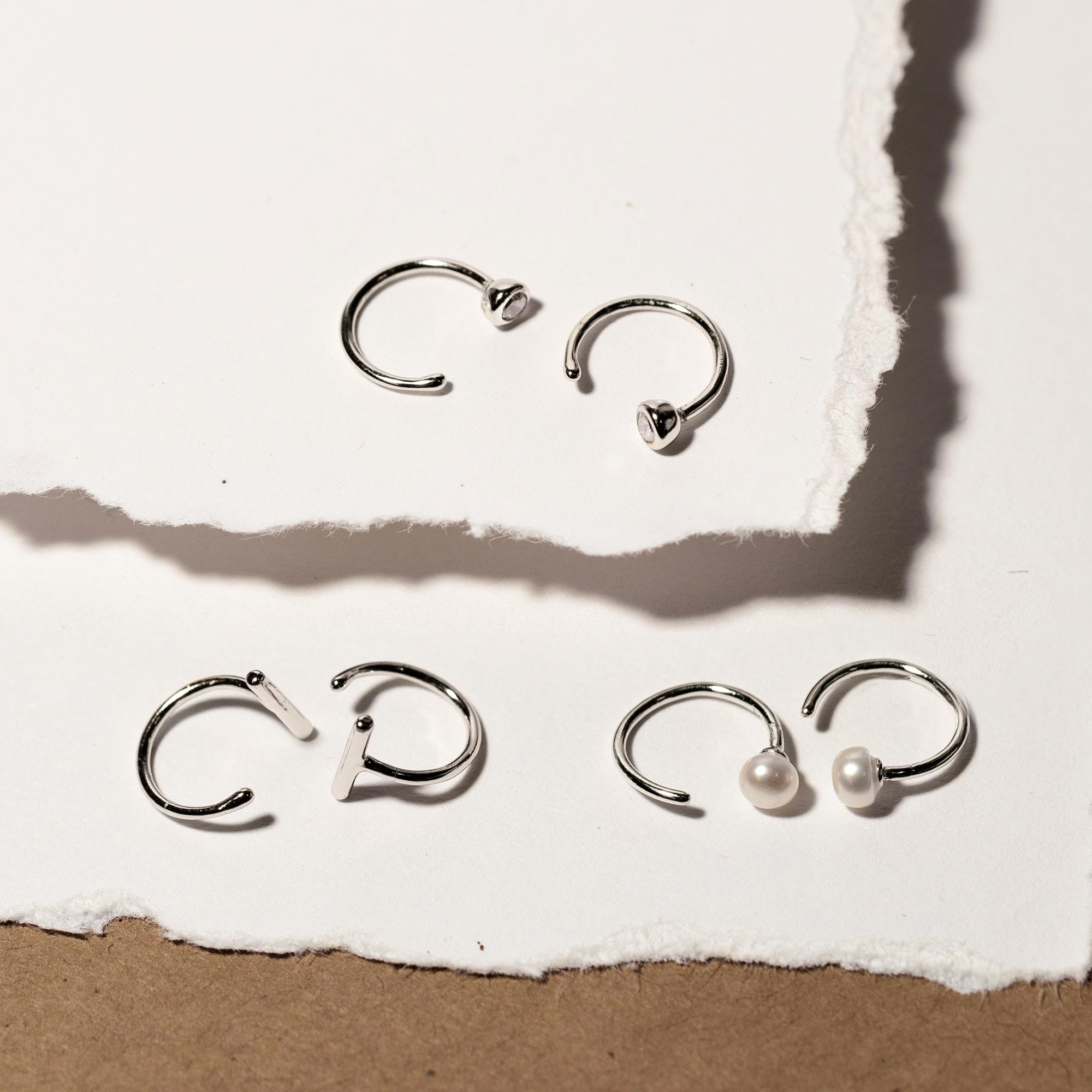Sterling silver open huggie hoops with white zirconia, shown with pearl and plain styles on paper