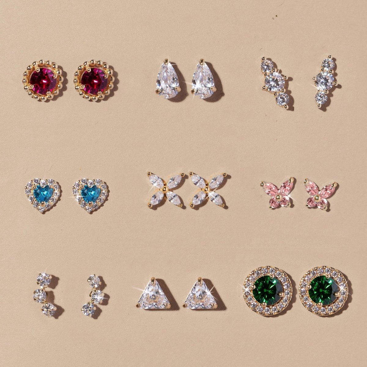 An assortment of gold studs in varied shapes with white, pink, blue and green stones, on a cream background