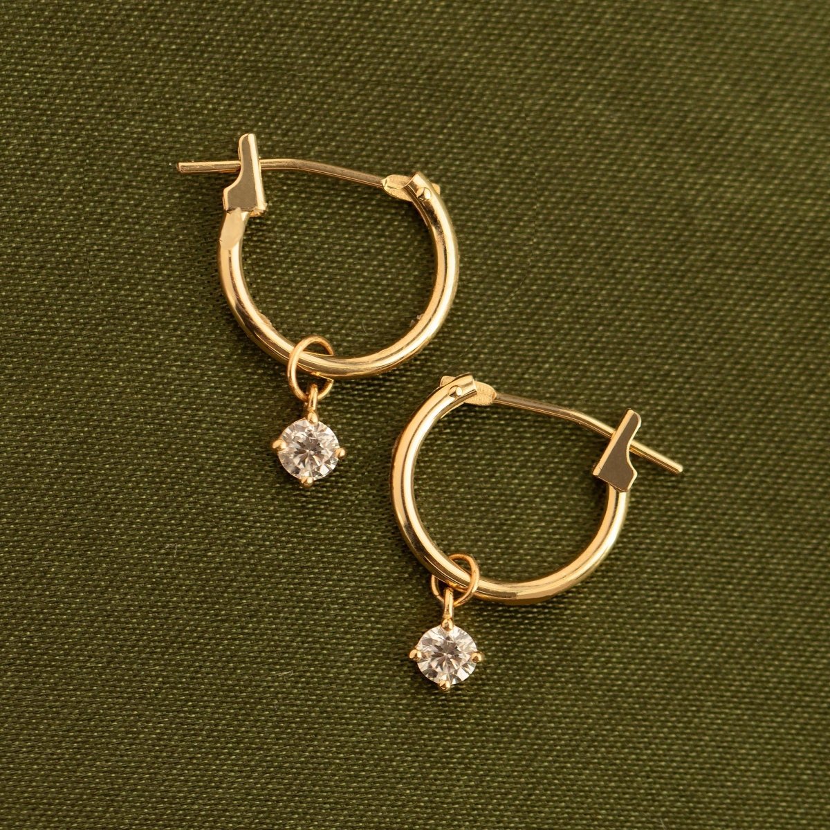 Pair of 14K gold huggie hoops with round clear moissanite charms, laid on olive fabric