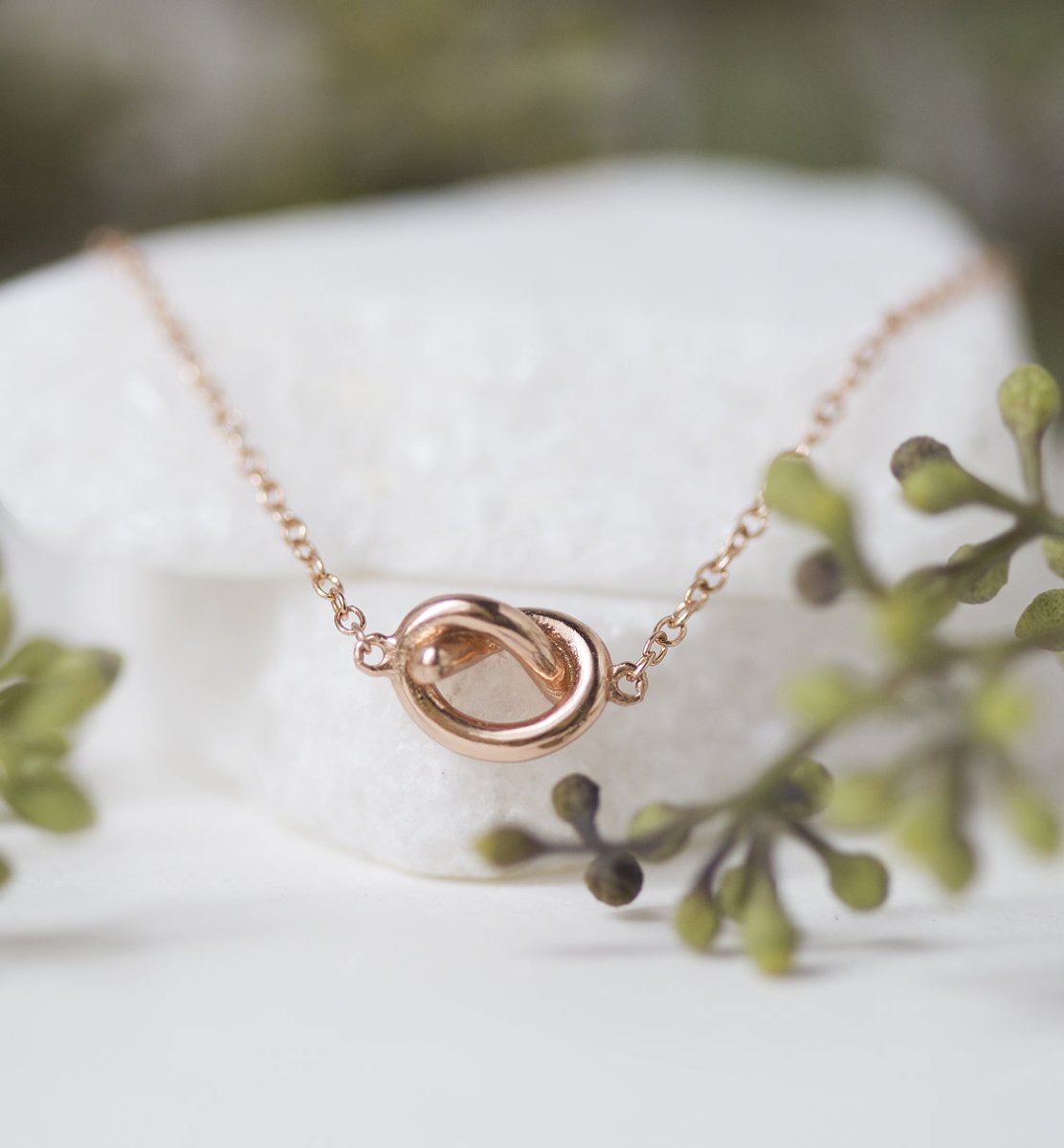 Rose gold vermeil love knot necklace close-up detail, shown against a white stone with greenery