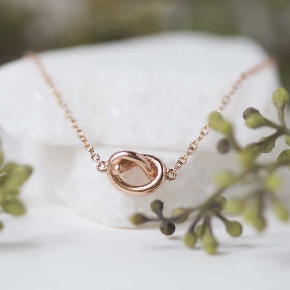 Rose gold vermeil love knot necklace close-up detail, shown against a white stone with greenery