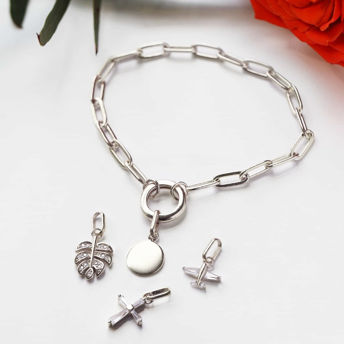 Sterling silver paperclip chain charm bracelet with clasp ring and loose charms beside a red rose
