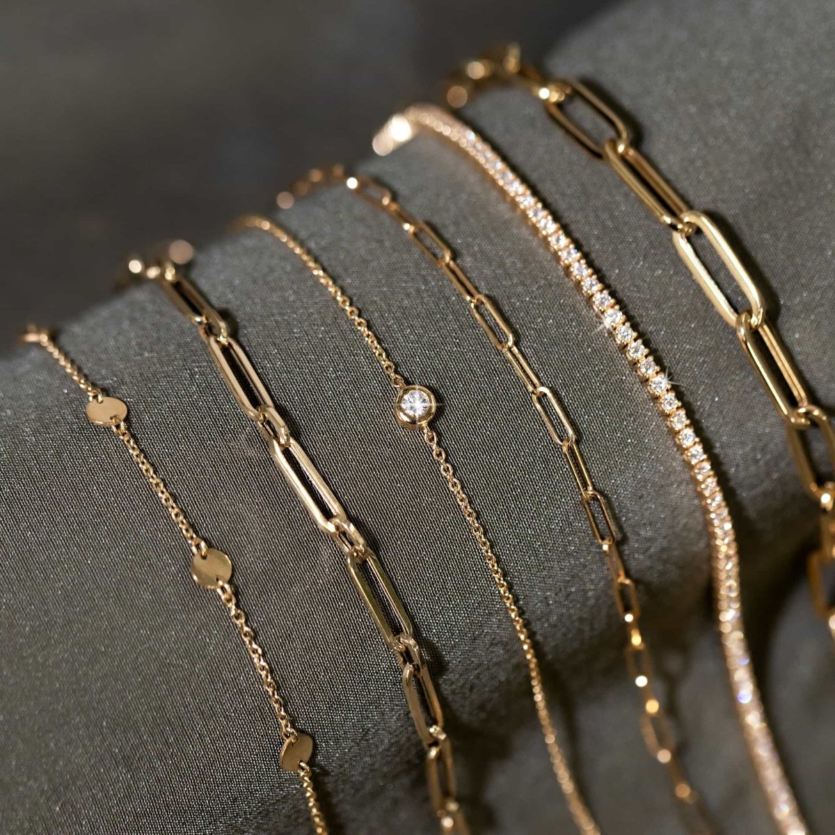 14K gold bezel diamond chain bracelet layered with gold bracelets on draped fabric