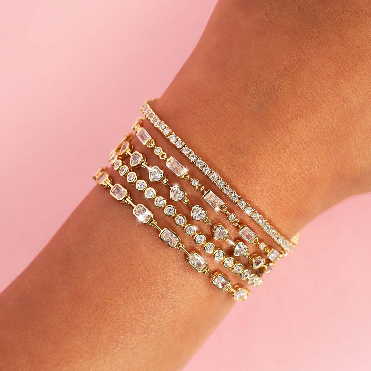 Gold tennis bracelets with mixed-cut clear stones, stacked on a model's wrist on pink