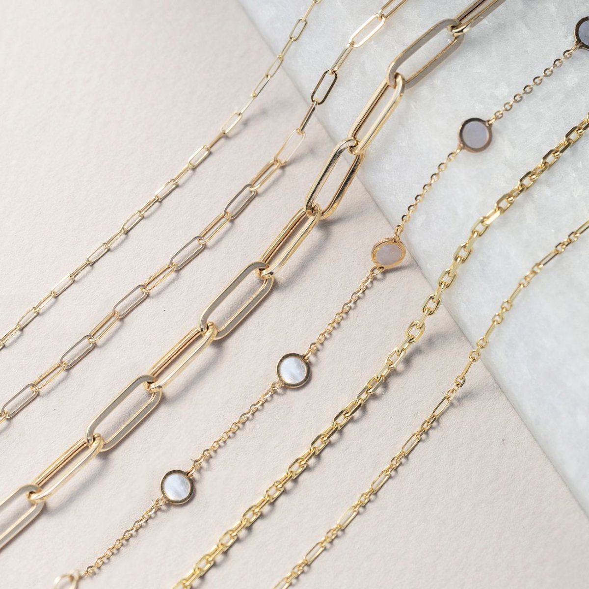 Assorted gold chain bracelets, including paperclip and stone-station styles, on white marble and cream