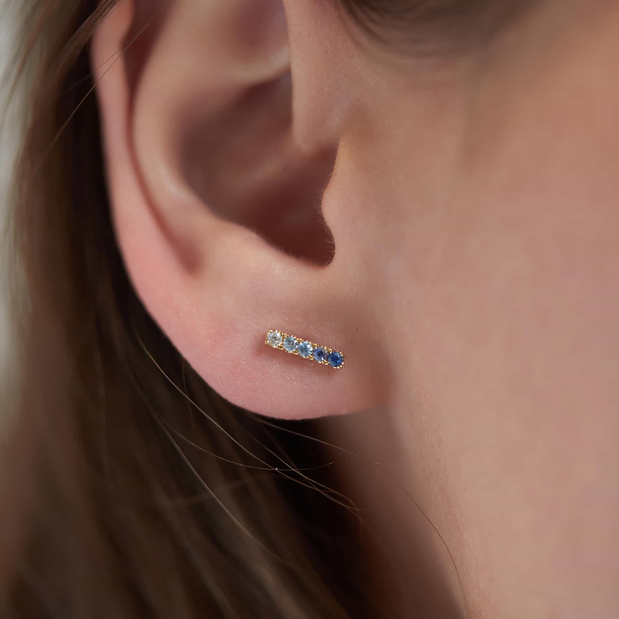 14K gold bar stud with ombre clear-to-blue sapphire stones, shown on a model's lobe