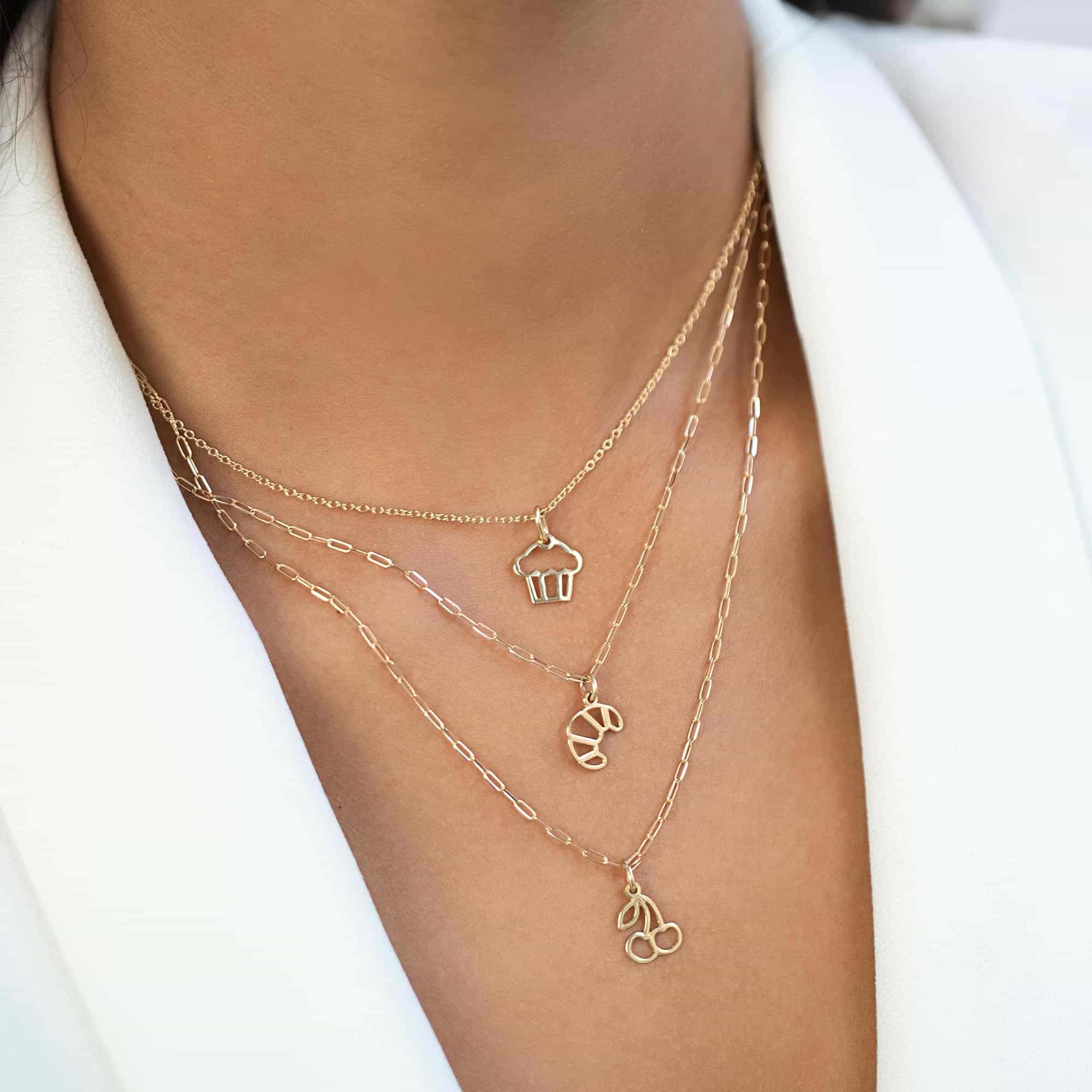 Layered gold necklaces with cupcake, croissant and cherry charm pendants, shown on a model's neckline