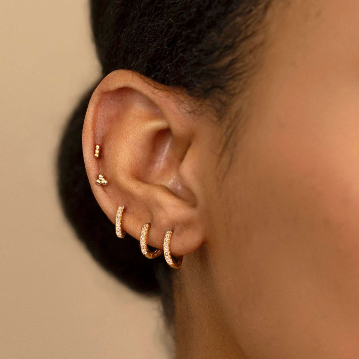 Gold vermeil tiny ball cluster studs shown on a model's upper lobe above pave huggie hoops