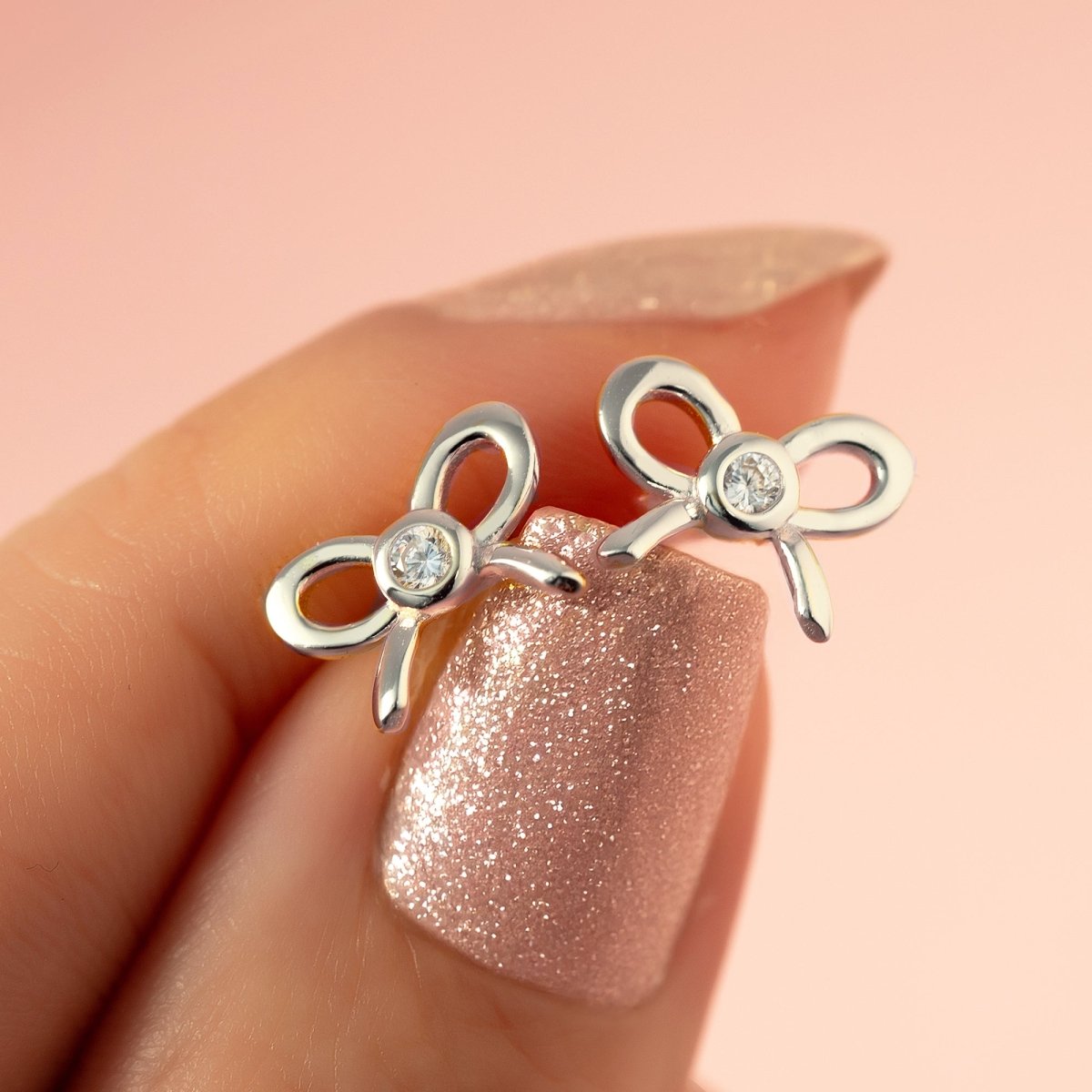 Sterling silver bow studs with clear bezel stones held against a fingertip, close-up detail