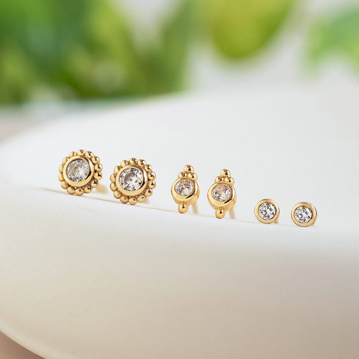 Three pairs of gold bezel studs with clear stones in graduated sizes, on a cream dish with greenery