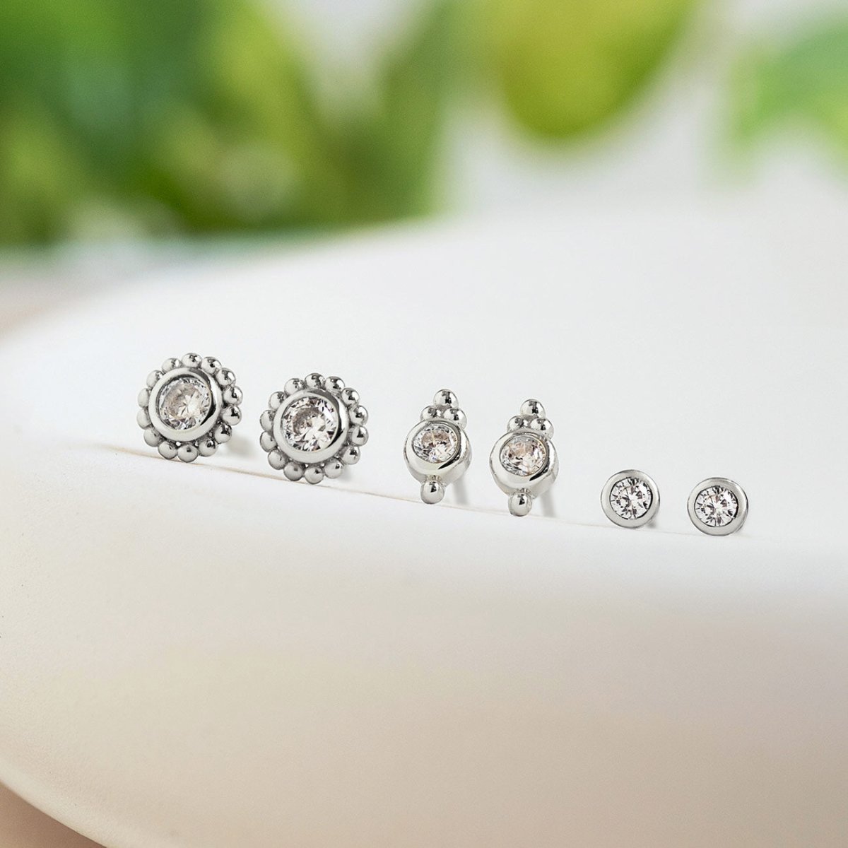 Three pairs of silver bezel studs with clear stones in graduated sizes, on a cream dish with greenery