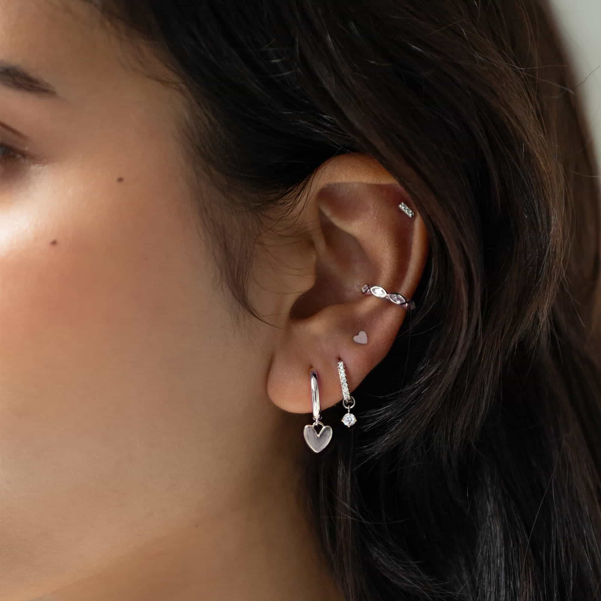 Close-up of woman's ear featuring 14K White Gold Tiny Gemstone Pavé Huggies Moissanite and Sterling Silver Heart Earrings.
