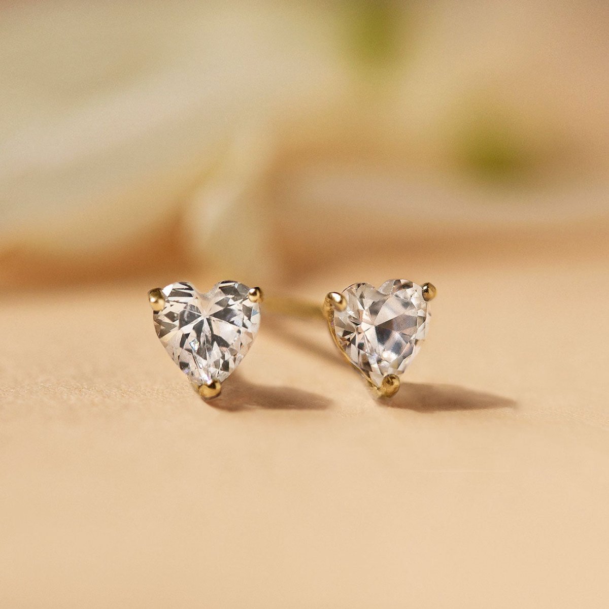 Gold vermeil heart studs with clear white zirconia stones, close-up detail on a cream surface