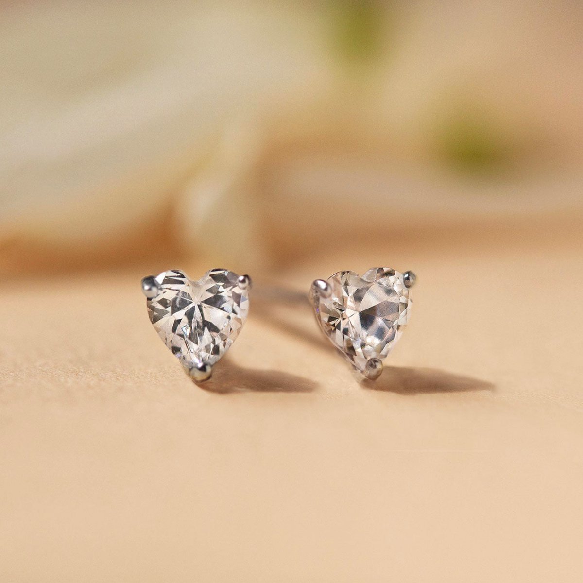 14K white gold heart-cut white zirconia stud earrings, close-up detail on a warm cream surface