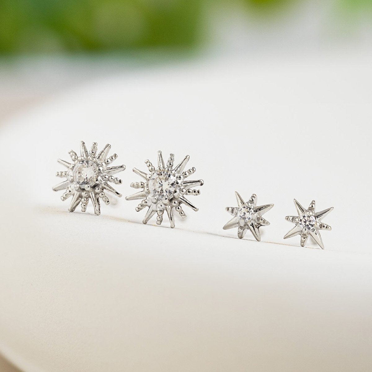 Sterling silver starburst studs with white zirconia, two sizes shown side by side on a cream background