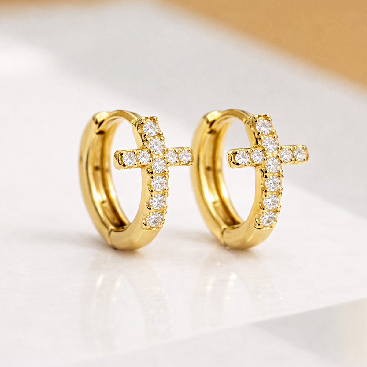 Gold vermeil cross huggie hoops with pavé clear crystals, shown angled on a cream surface