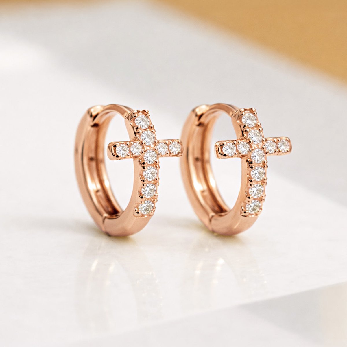Rose gold vermeil cross huggie hoops with pavé clear crystals, shown angled on a cream surface