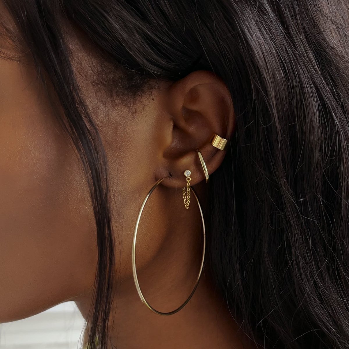 Close-up on woman's ear featuring Gold Vermeil Thin Large Hoops, Suspender Earrings, Ear, Cuff, and a Gold Chain Earring.