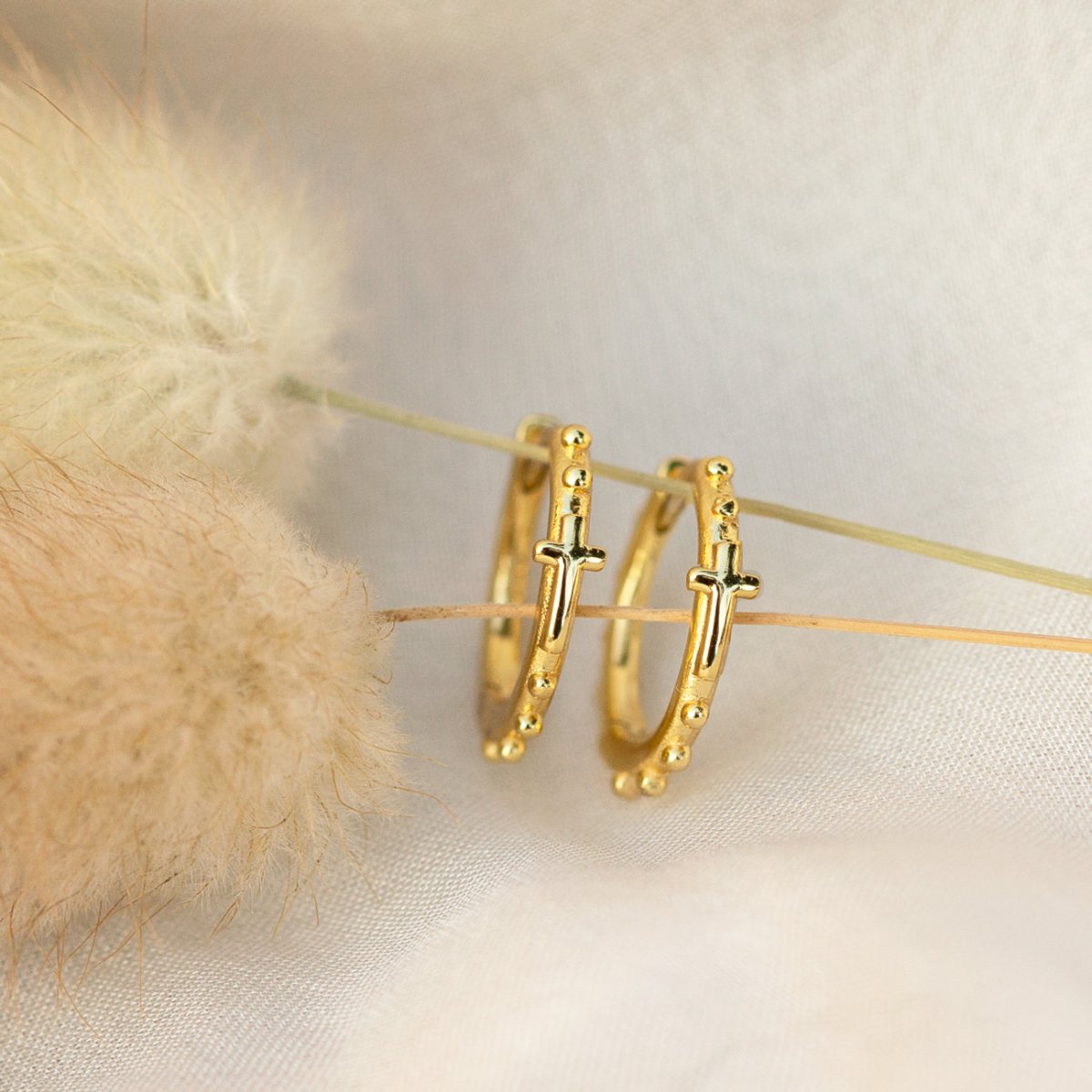 Gold vermeil beaded cross huggie hoops shown propped against dried flowers on cream fabric