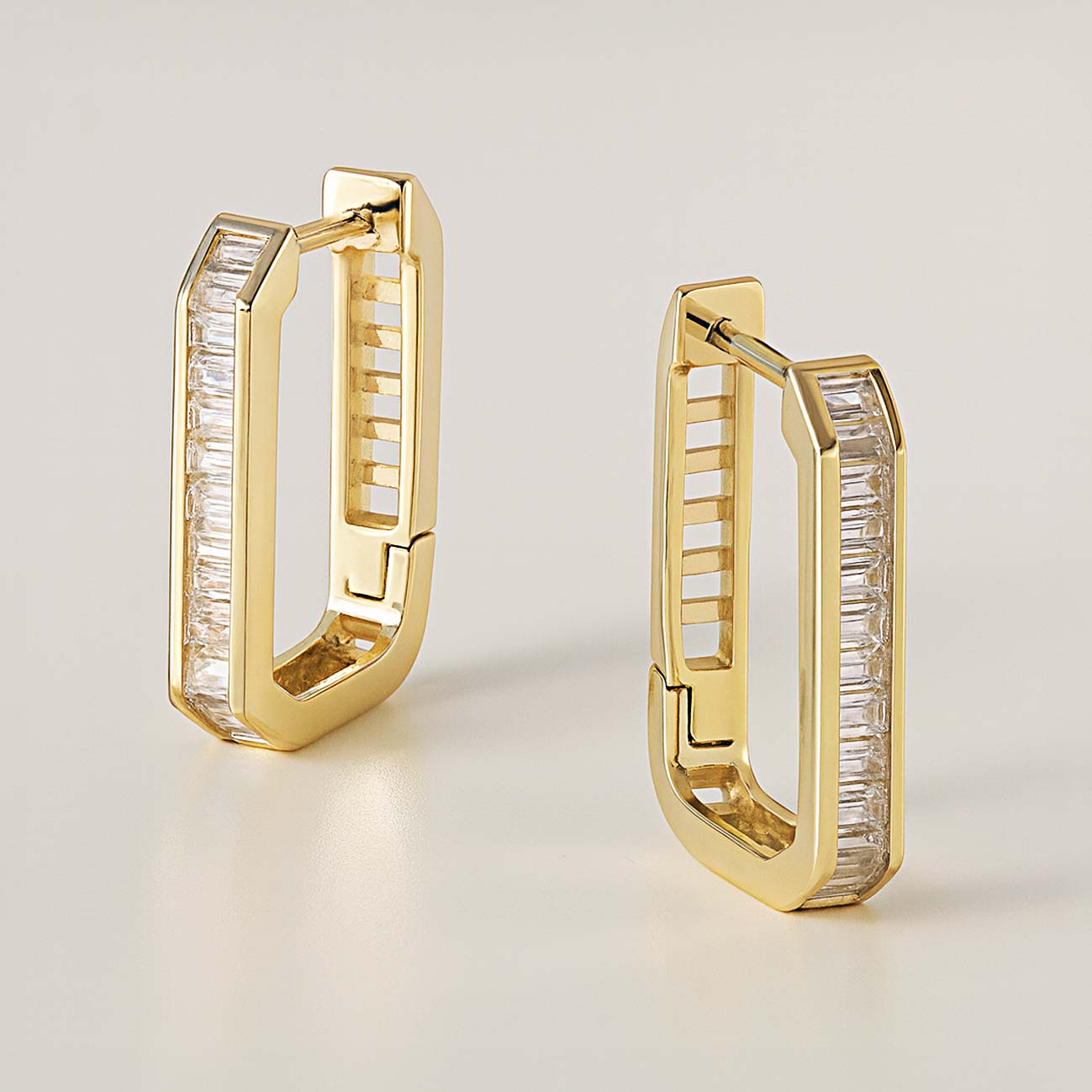 Gold vermeil channel-set baguette oval huggie hoops with clear stones, pair shown standing on a cream background