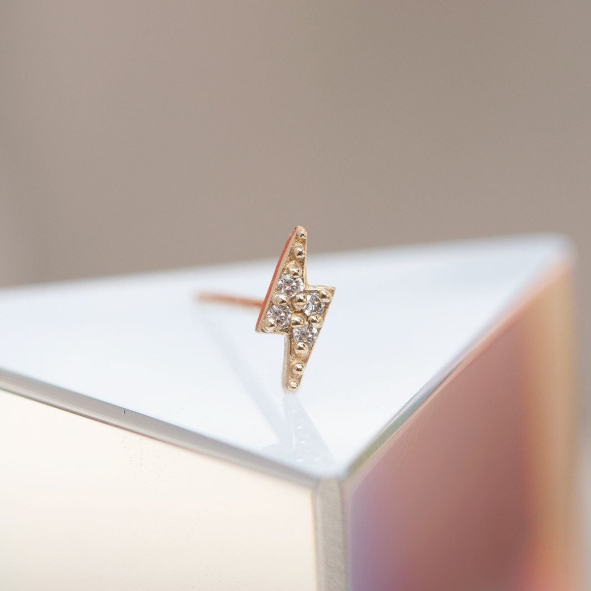 14K gold diamond pavé lightning bolt stud resting on a white block, soft light