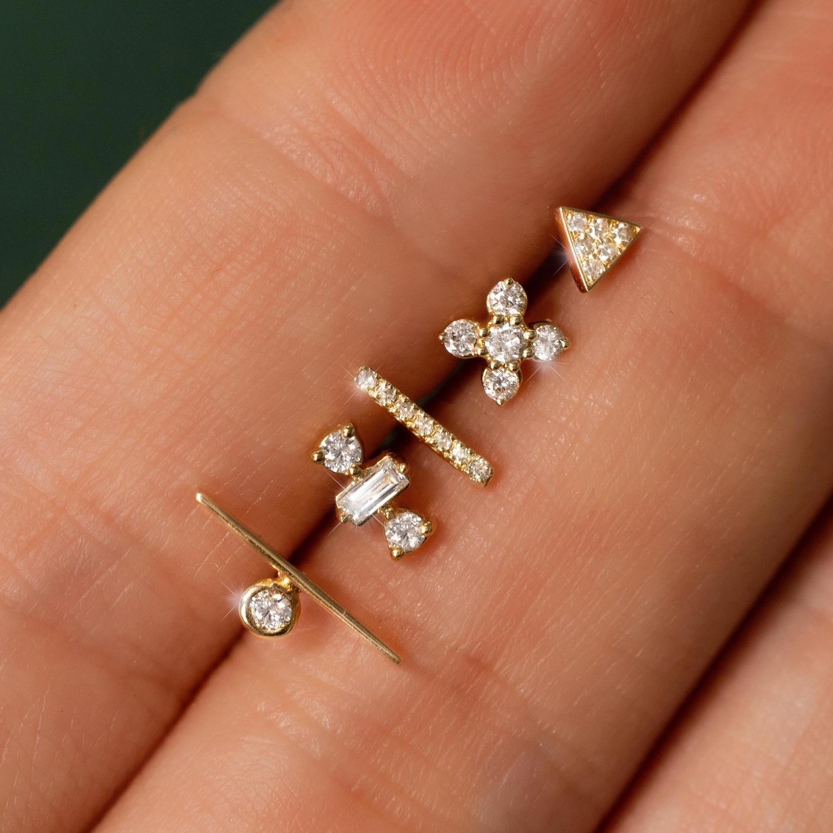 Close-up of 14K gold diamond studs in varied shapes held on a fingertip, with a baguette triad stud
