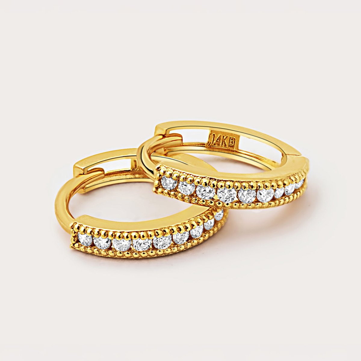 Two 14K gold milgrain diamond huggie hoops resting together, close-up detail on a cream background