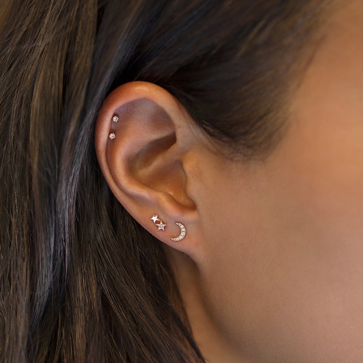 Gold star, moon and clear-stone studs shown on a model's lobe and helix piercings