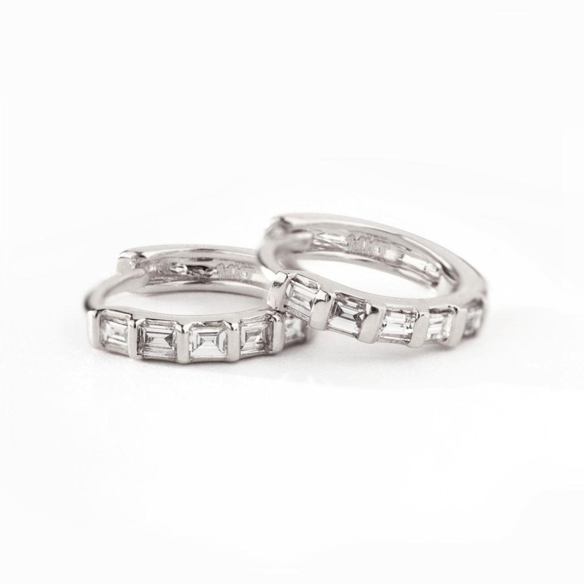 Pair of 14K white gold baguette diamond huggie hoops shown resting on a white surface