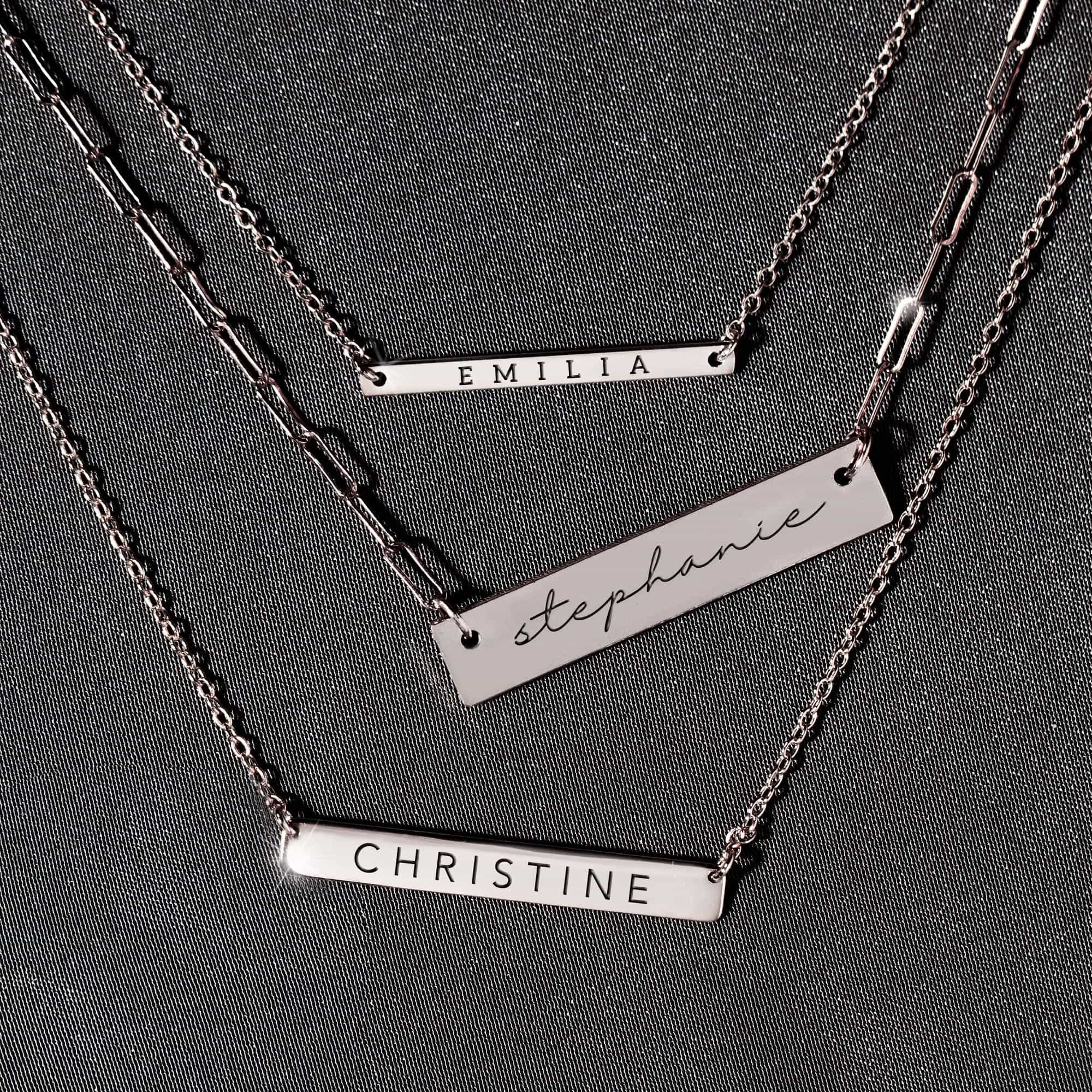 Sterling silver engravable bar necklaces engraved with names, layered on a gray backdrop