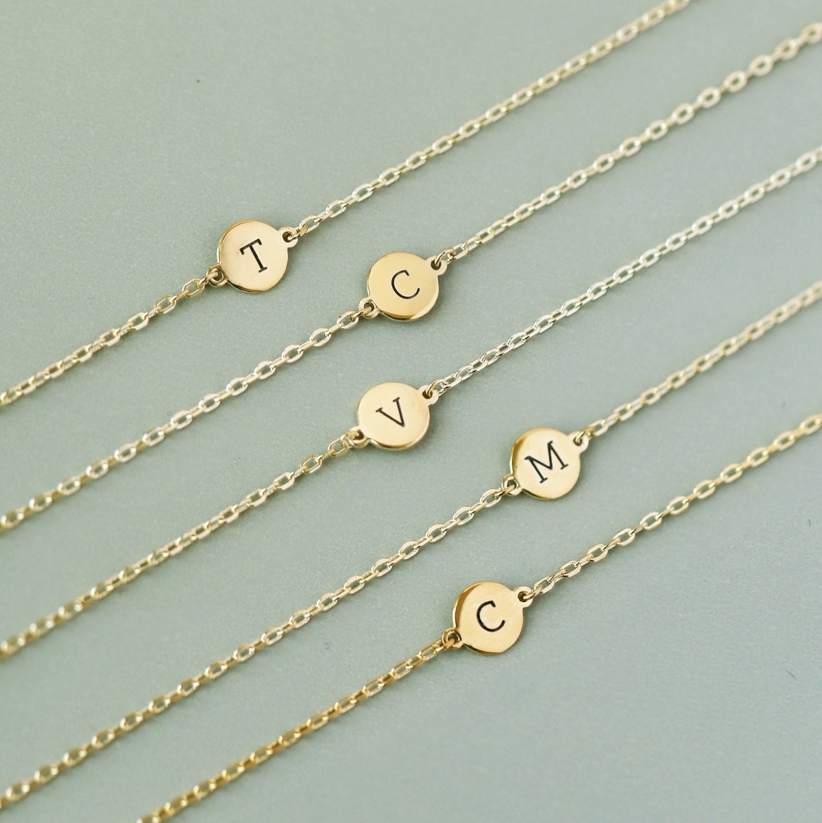Gold vermeil disc bracelets engraved with initials, laid in a row on sage green
