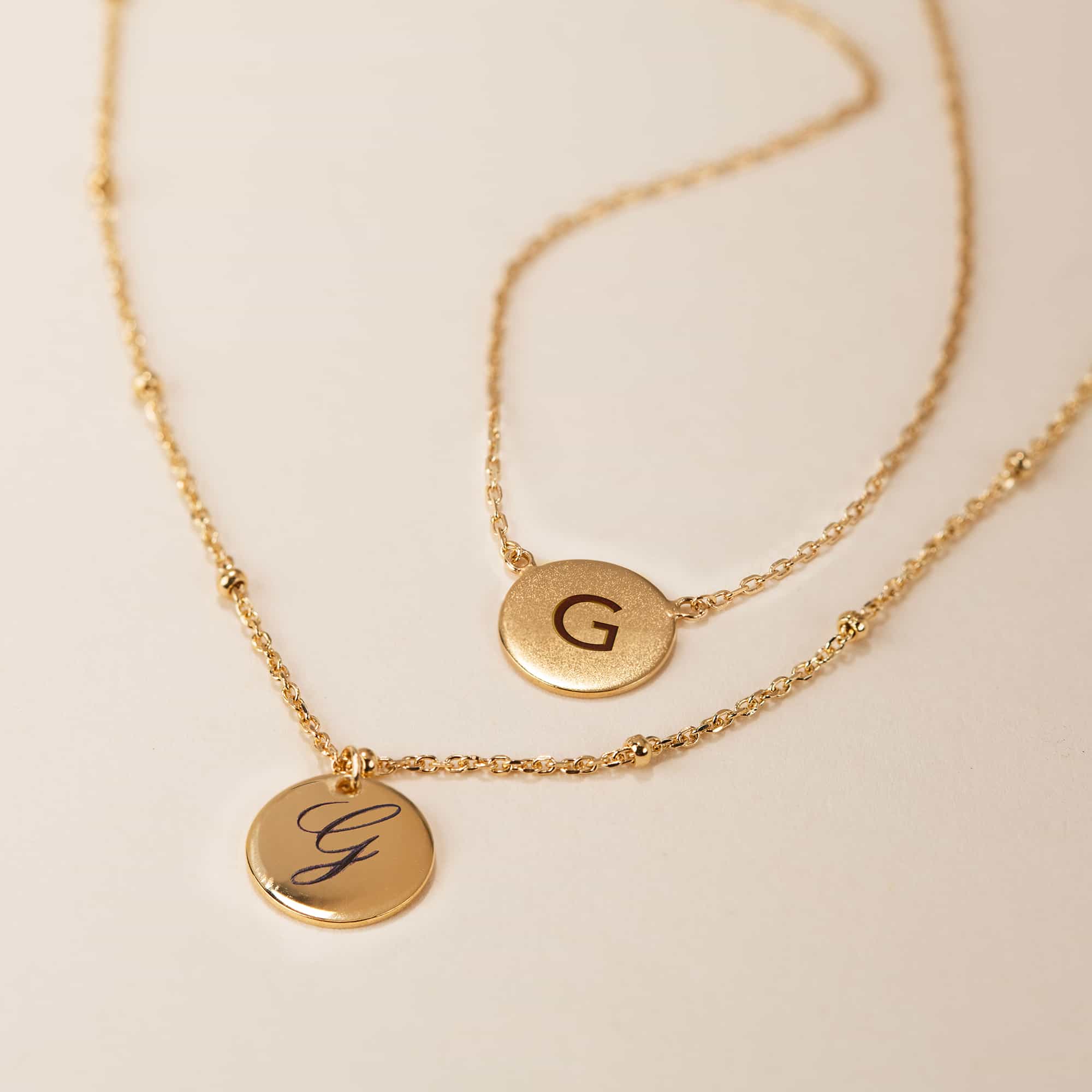 Two gold vermeil initial disc necklaces, engraved with an initial in block and script, on a cream background
