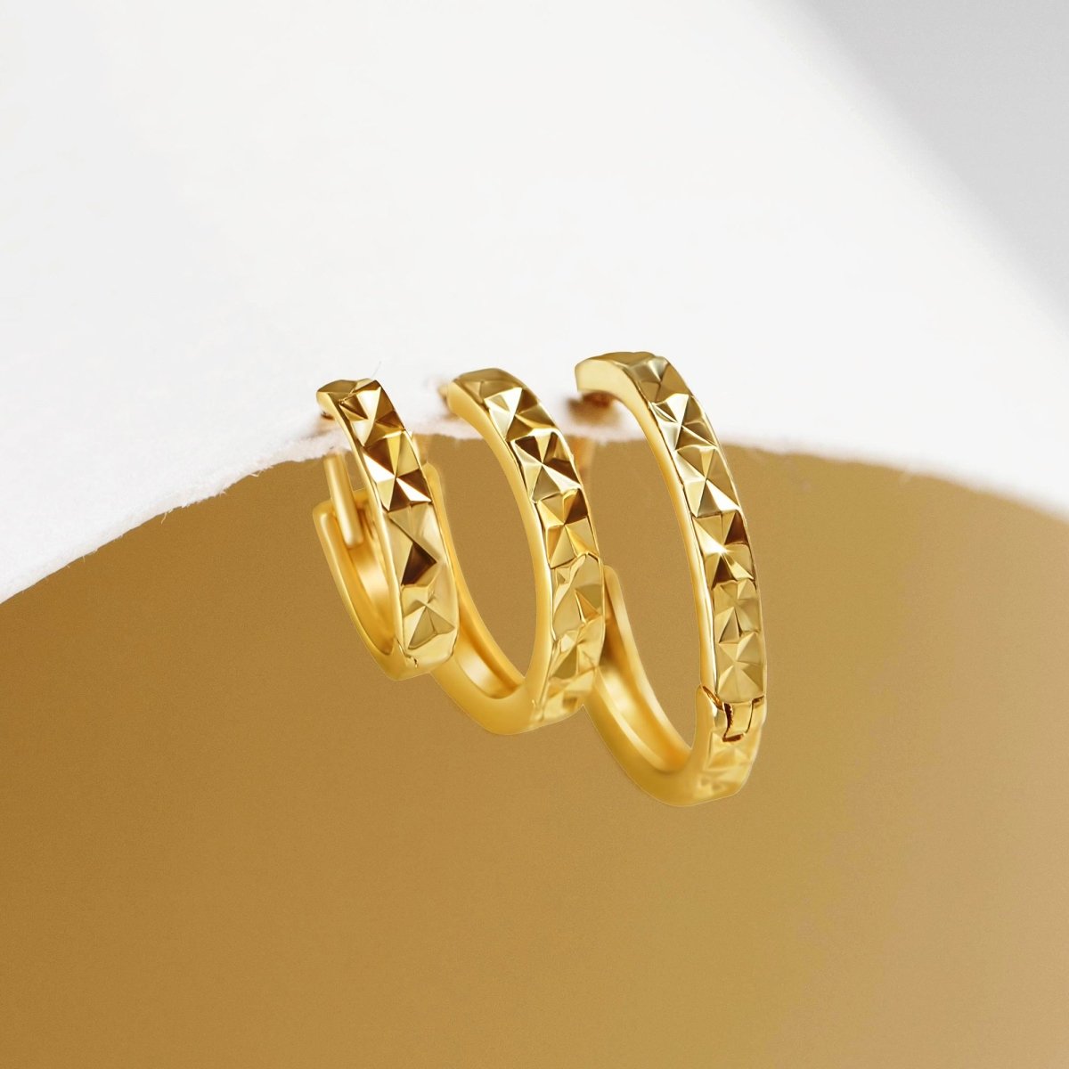 14K gold diamond-cut huggie hoops with a faceted texture, propped on a gold surface