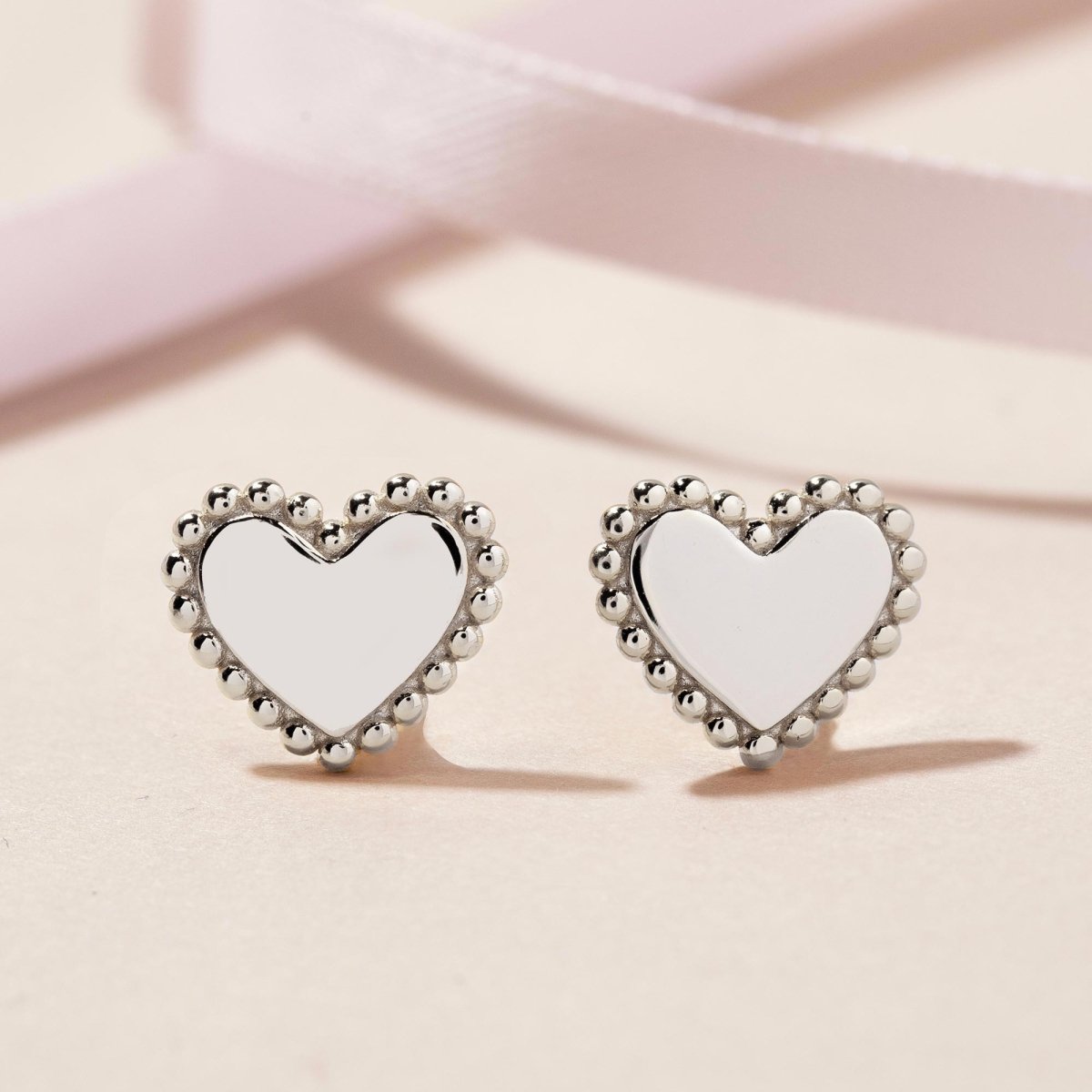 Sterling silver heart stud earrings with beaded edges, on a pink ribbon background