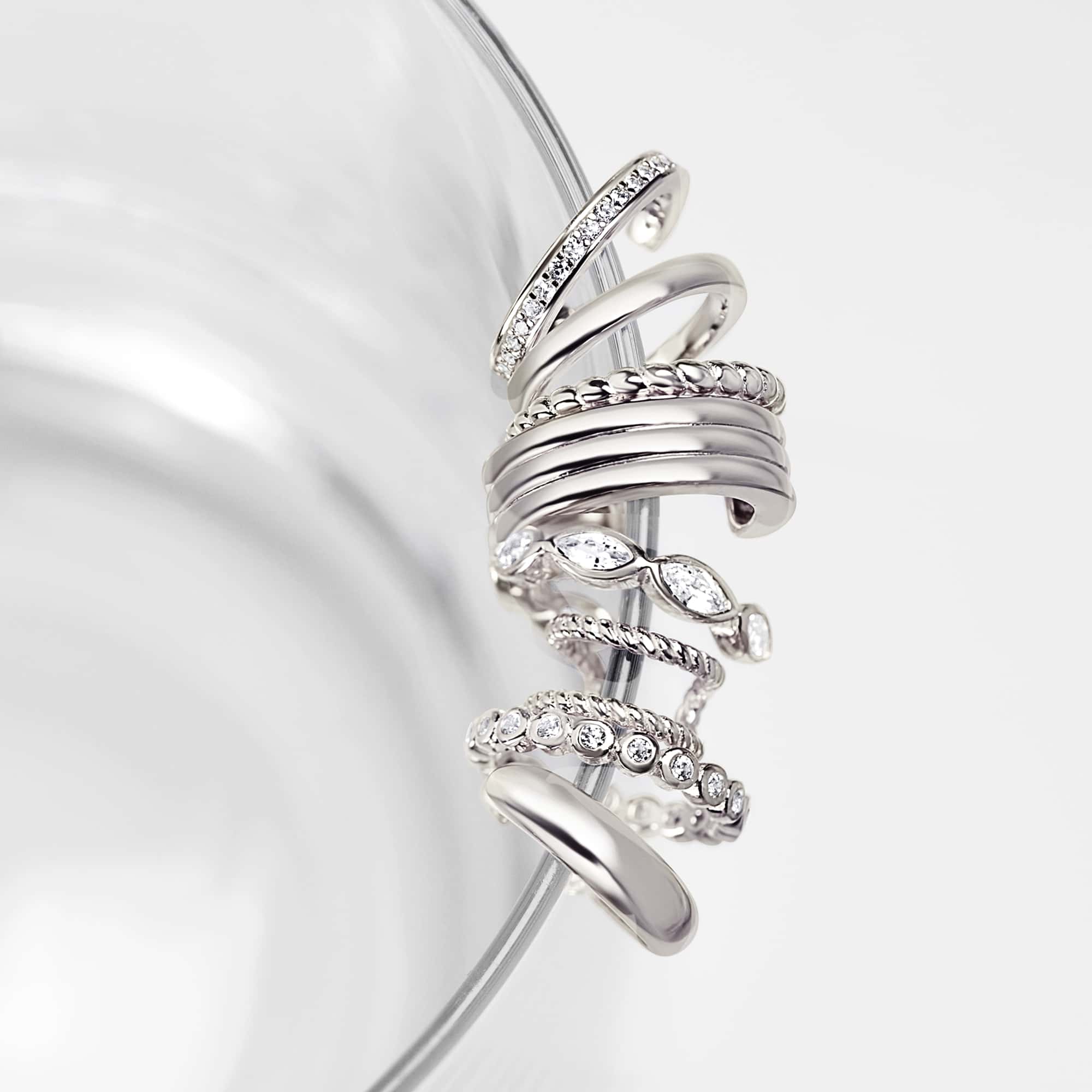 Assortment of silver ear cuffs with clear stones, rope and plain styles, coiled on a clear glass rim