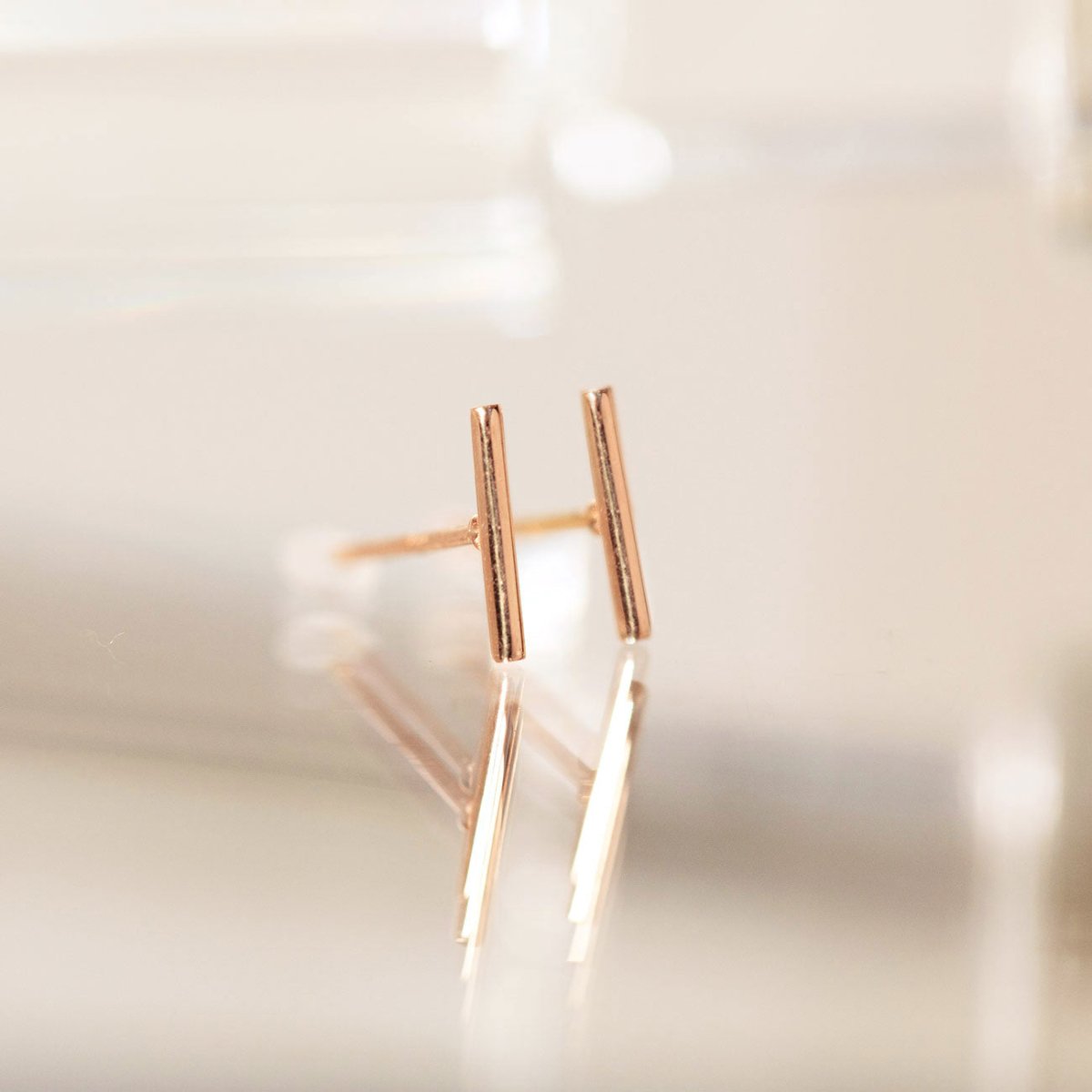 14K rose gold thin bar stud earrings standing upright, close-up on a soft white surface