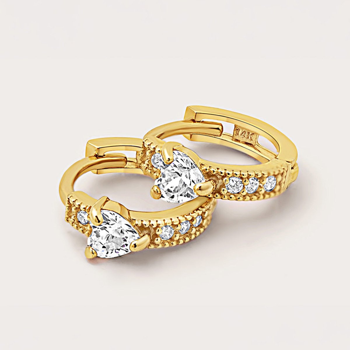 14K gold heart huggie hoops with white zirconia stones, close-up detail on a cream background