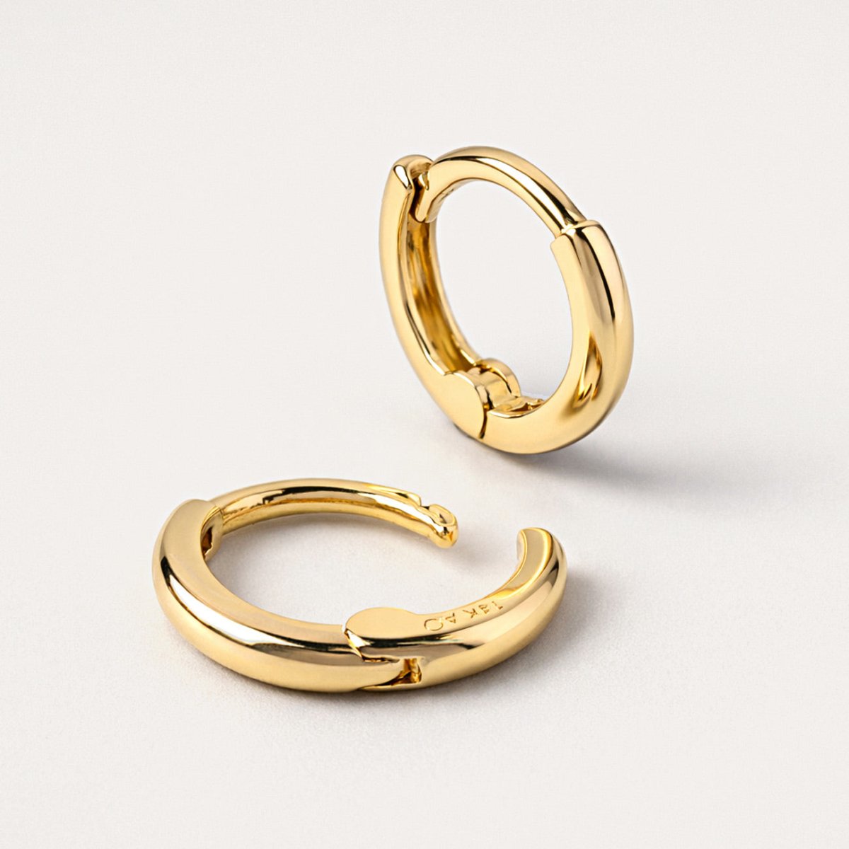14K gold mini huggie hoops close-up detail showing the smooth band and hinged closure, on a cream background